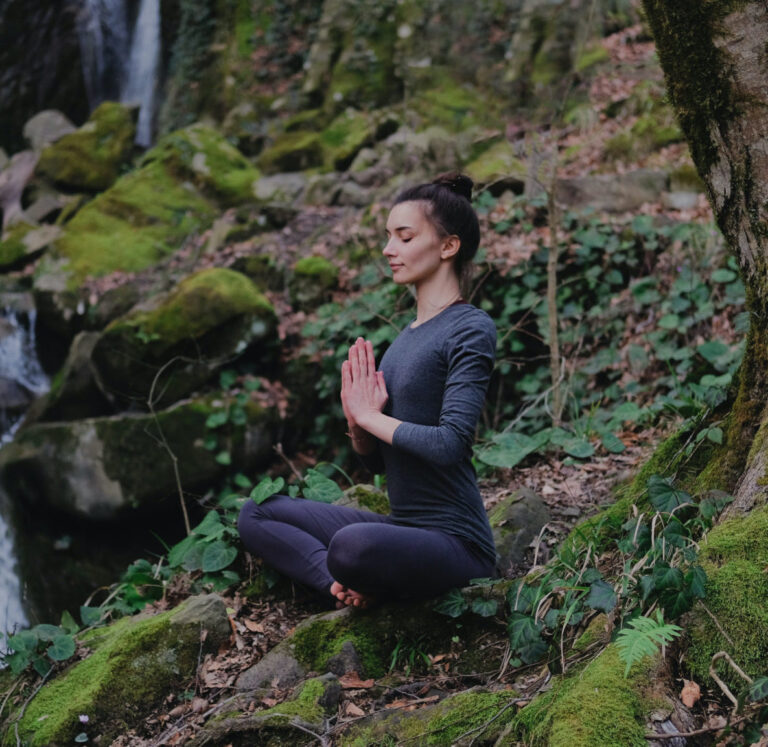 Young slim woman practicing yoga outdoors in moss forest on background of waterfall. Unity with nature concept. Girl meditates sitting.